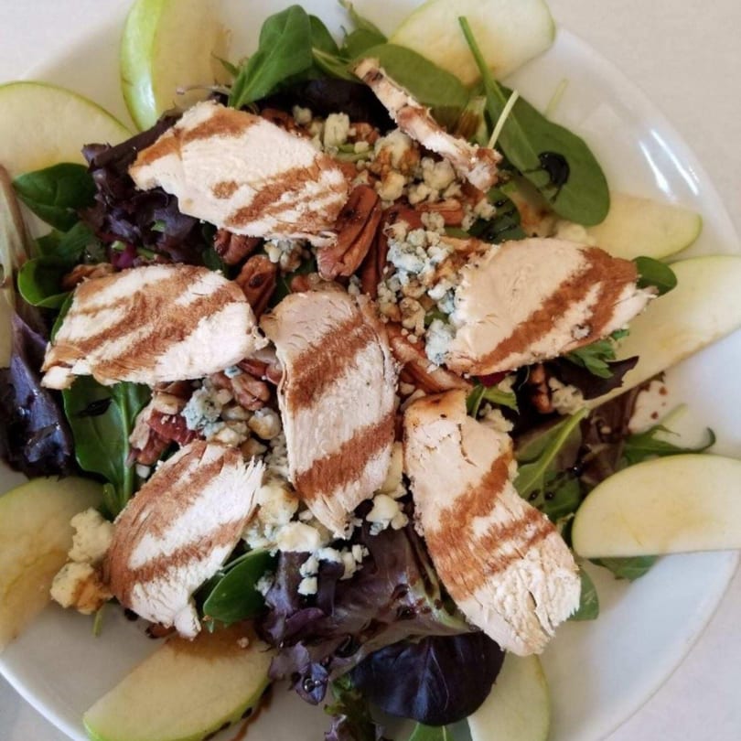 A vibrant salad with grilled chicken, blue cheese, pecans, and green apple slices on a white plate.