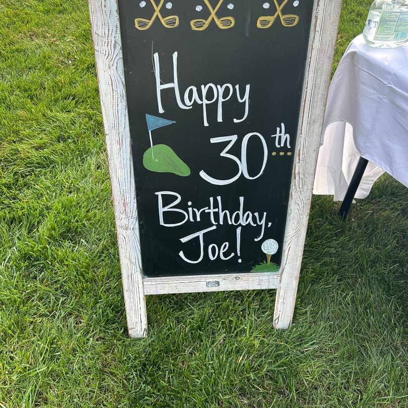 Chalkboard sign on grass with "Happy 30th Birthday, Joe!" and golf-themed decorations.