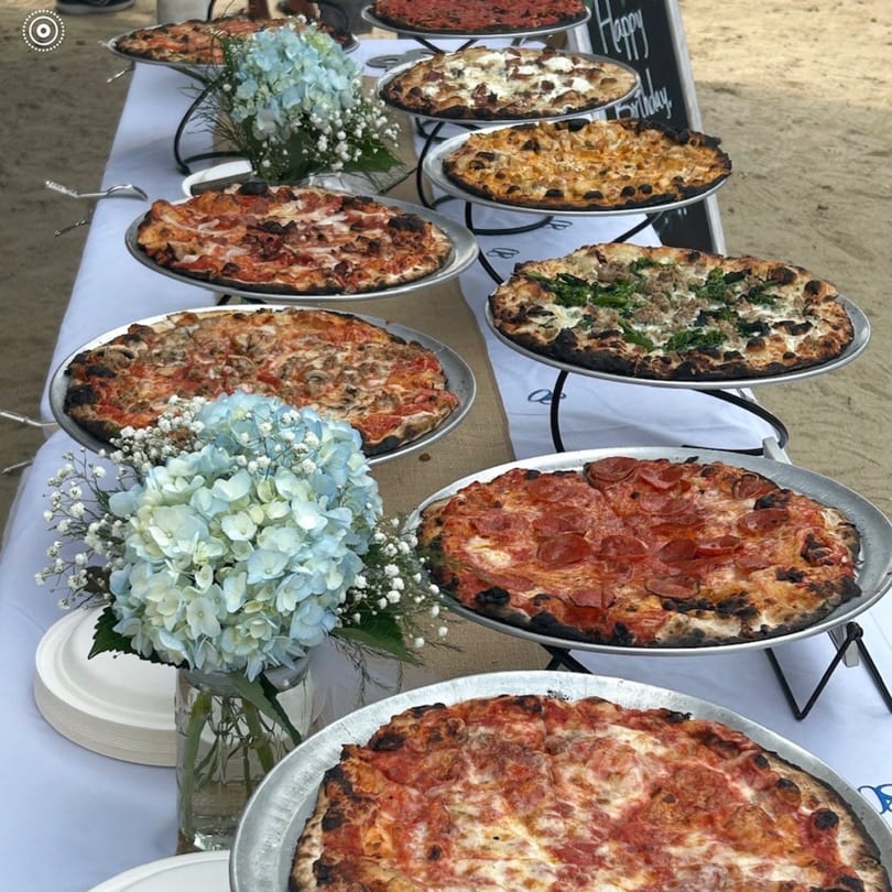 A long table covered with a white cloth, displaying many pizzas on metal stands, with blue floral arrangements.