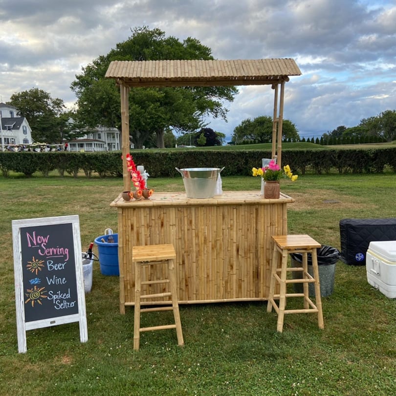 A bamboo bar with two stools, a "Now Serving" sign, and coolers on a grassy lawn.