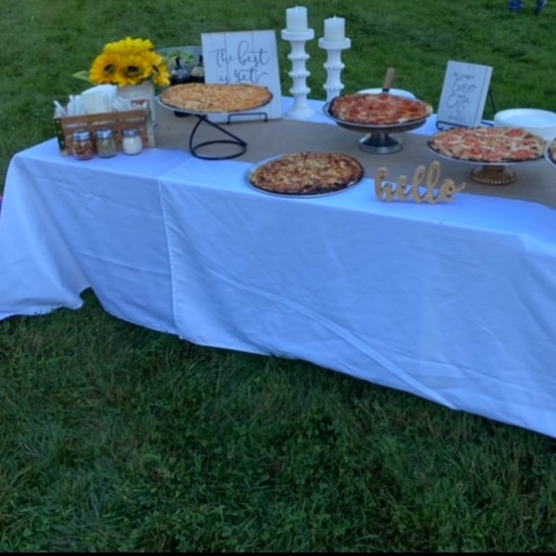 Outdoor buffet table with multiple pizzas, sunflowers, and decorative candles on a white tablecloth.