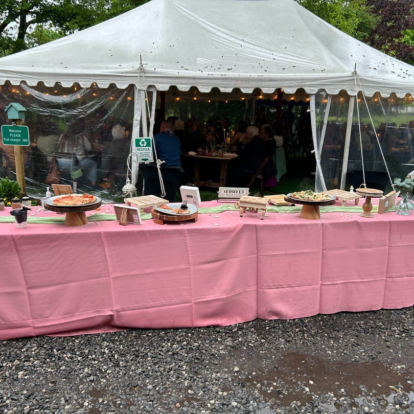 A pink-covered buffet table with pizzas and other dishes outside a white tent with diners.
