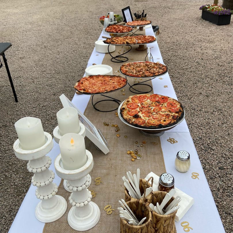 A long outdoor table laden with various Marco Pizzeria pizzas, candles, and serving supplies.