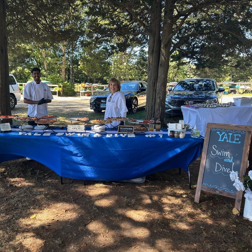 Two Marco Pizzeria chefs stand behind a blue table laden with pizzas and Italian food outdoors.