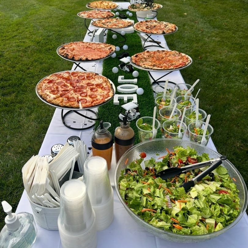 A long table with pizzas, salads, and golf-themed decorations set up outdoors on grass.