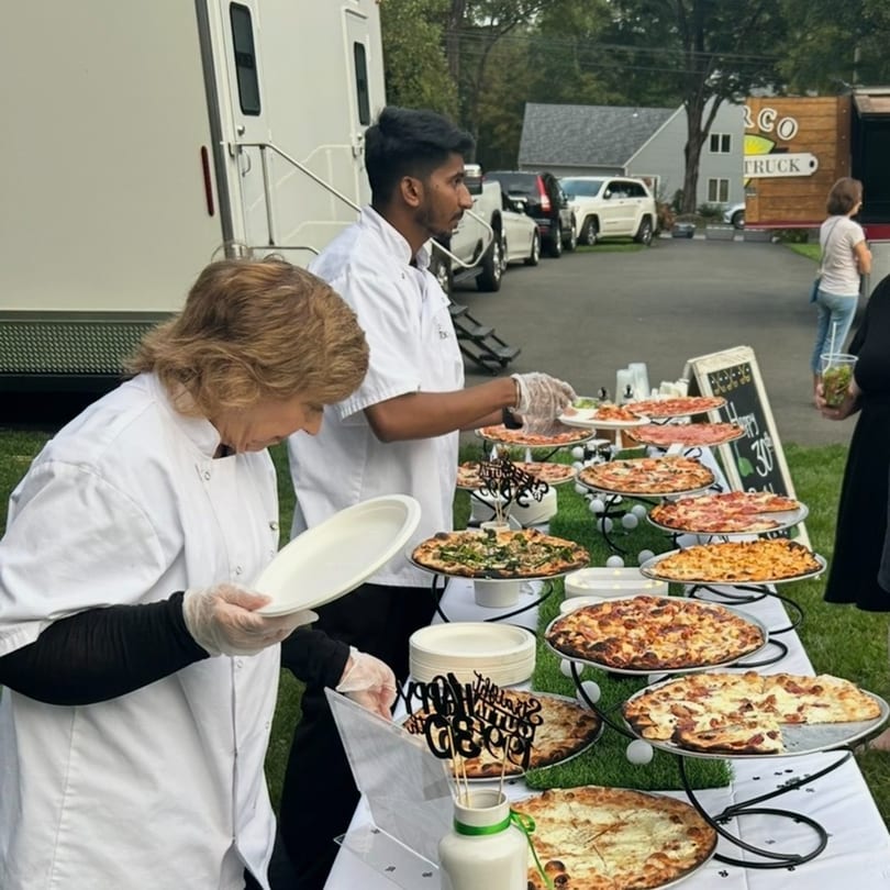 Chefs serving various Marco Pizzeria pizzas from a tiered display outdoors at an event.