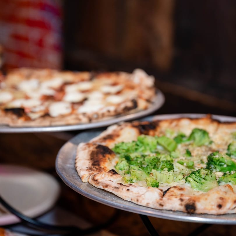 Two freshly baked Marco Pizzeria pizzas, one with broccoli and the other with white toppings.