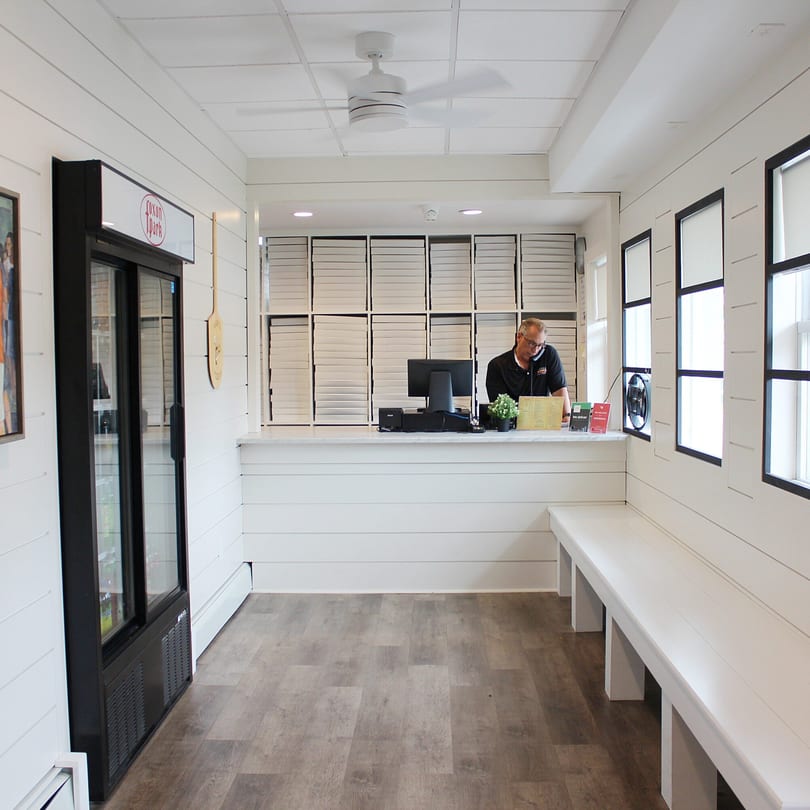 A Marco Pizzeria employee stands behind a counter with pizza boxes, a drink cooler, and benches.