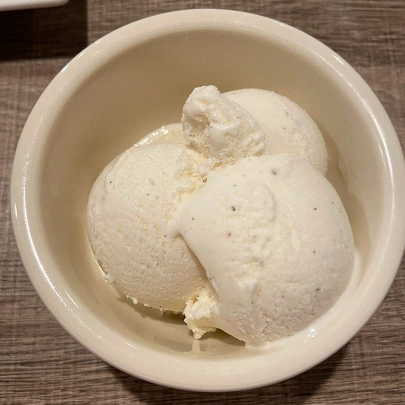Three scoops of creamy vanilla bean gelato in a white bowl on a wooden table.
