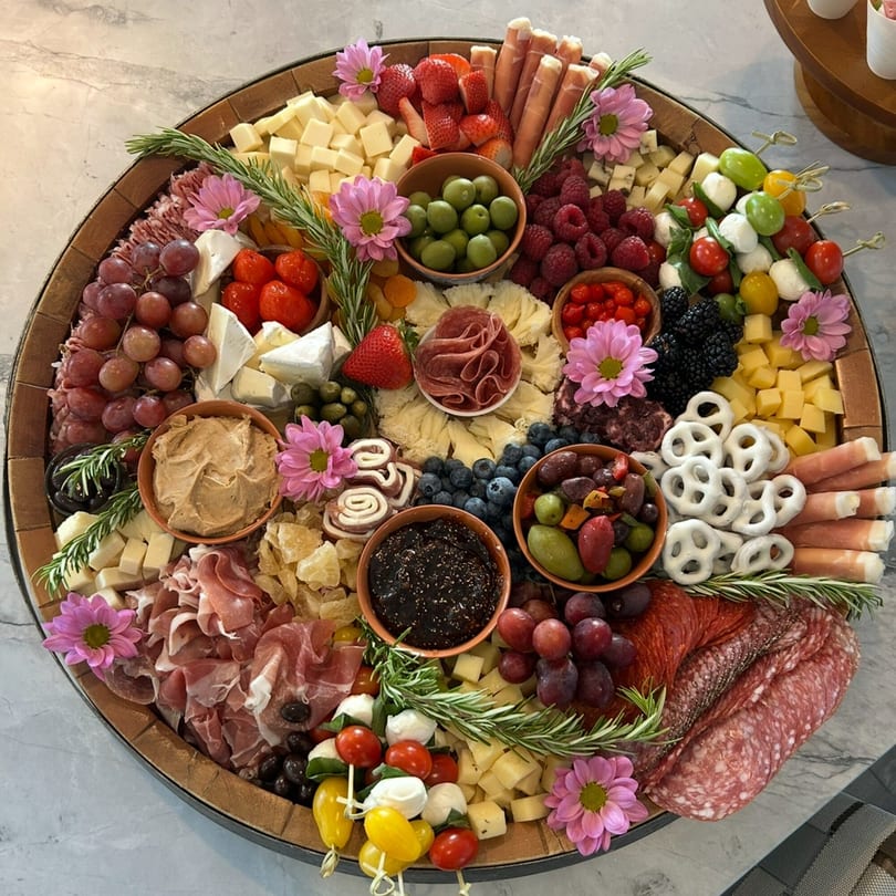 A large, round charcuterie board overflowing with Italian meats, cheeses, fruits, olives, and pretzels.