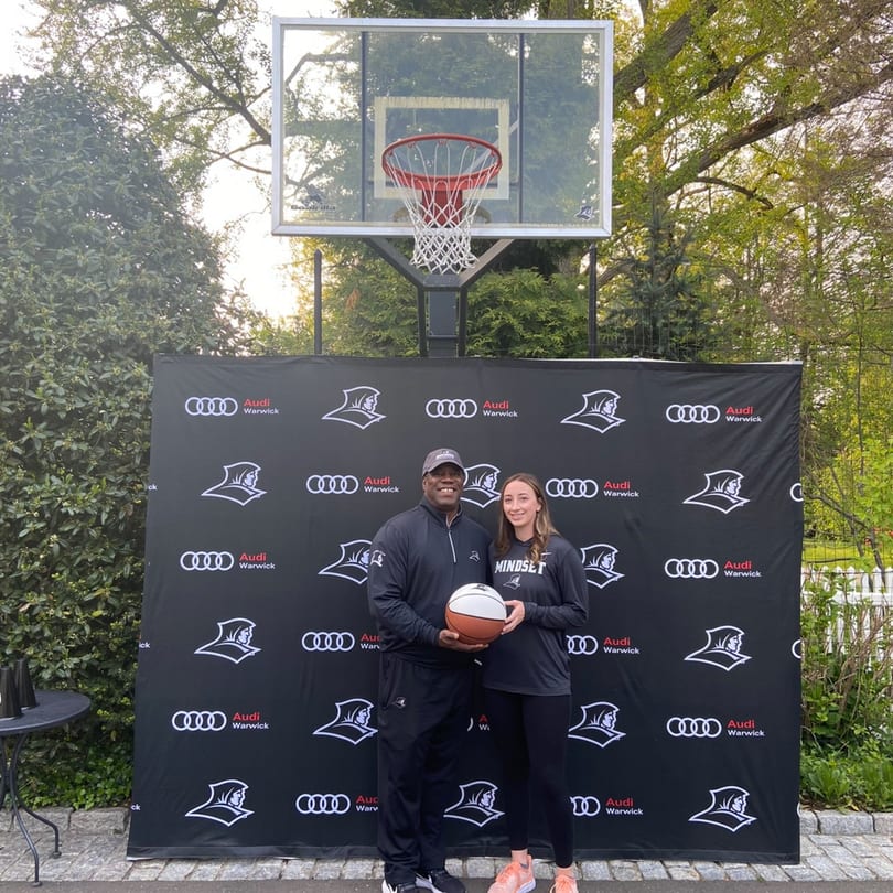 Two people holding a basketball in front of an Audi Warwick banner and a basketball hoop.