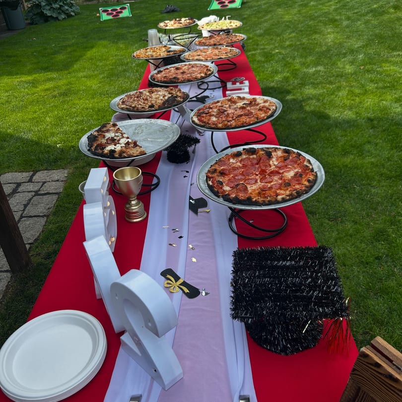 A long red table with a white runner holds multiple pizzas on stands, set on a green lawn.