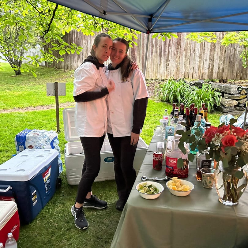 Two women in chef coats pose next to a bar setup with drinks and coolers outdoors.