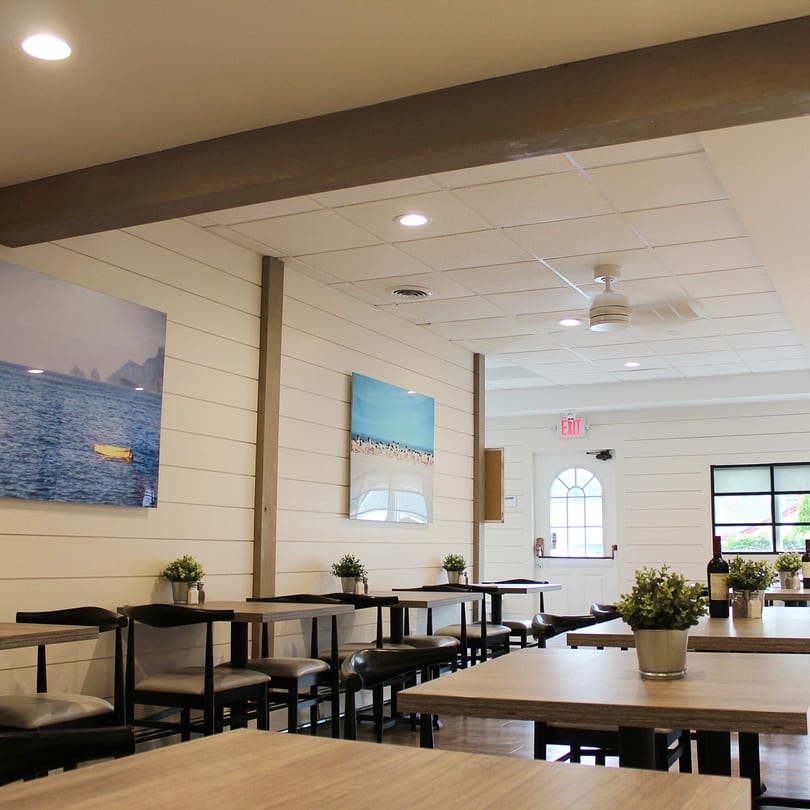 Bright, empty dining room at Marco Pizzeria with light wood tables, dark chairs, and nautical wall art.