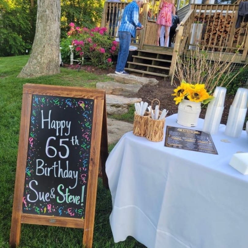 A chalkboard sign reads "Happy 65th Birthday Sue & Steve!" next to a white-clothed table with sunflowers.