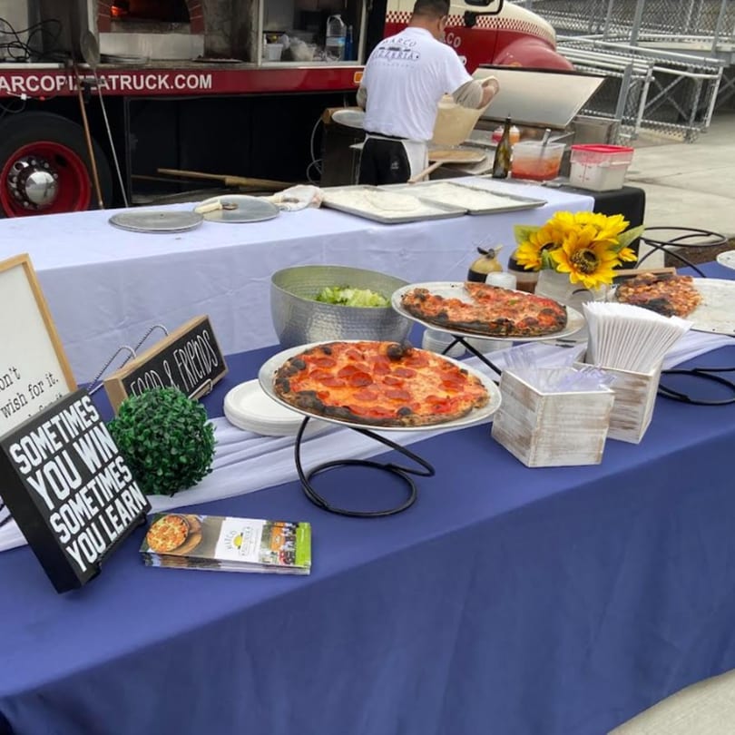 Marco Pizzeria food truck serving fresh pizzas and salad on a blue and white draped table.
