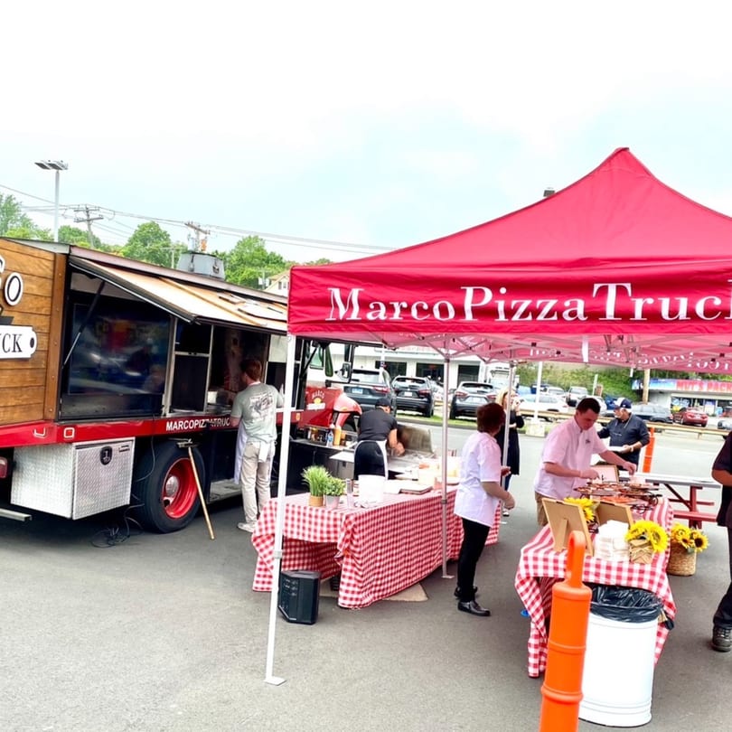 Marco Pizza Truck and red tent with checkered tables and people serving food outdoors.