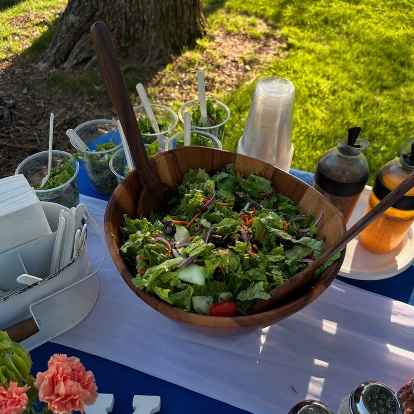 Large wooden bowl of fresh garden salad with dressing bottles and individual servings on an outdoor table.