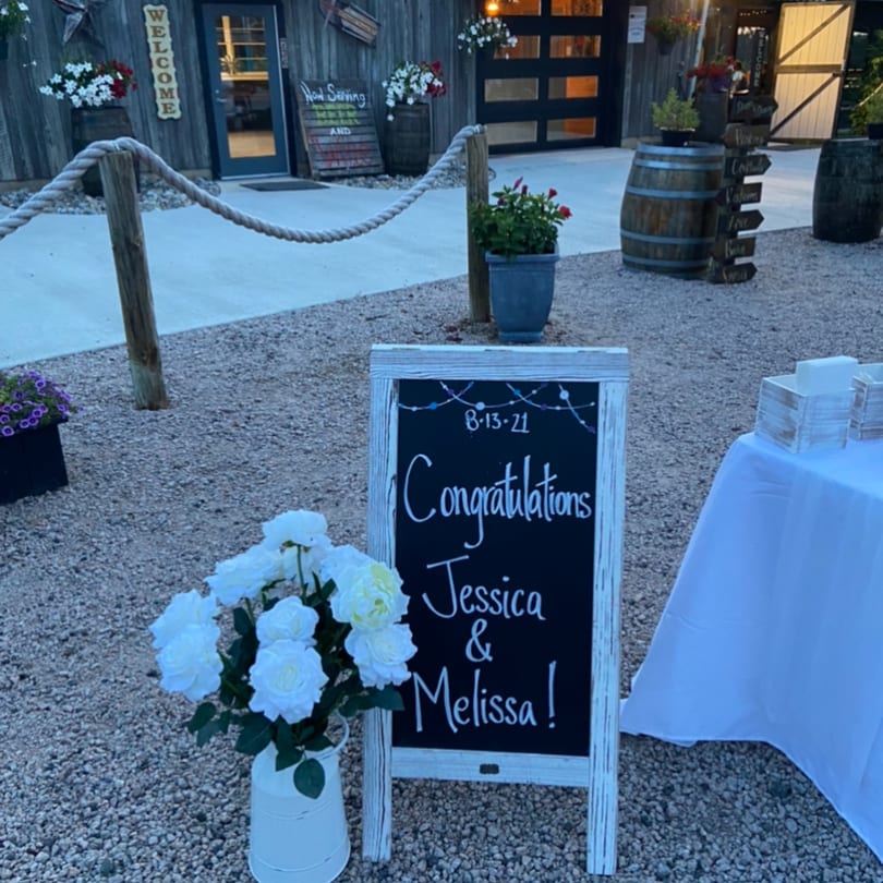 A chalkboard sign on a gravel path reads "Congratulations Jessica & Melissa!" with white roses beside it.