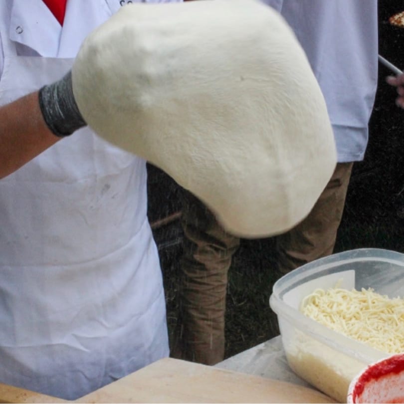 A chef in a white apron and black gloves stretches pizza dough, with shredded cheese nearby.