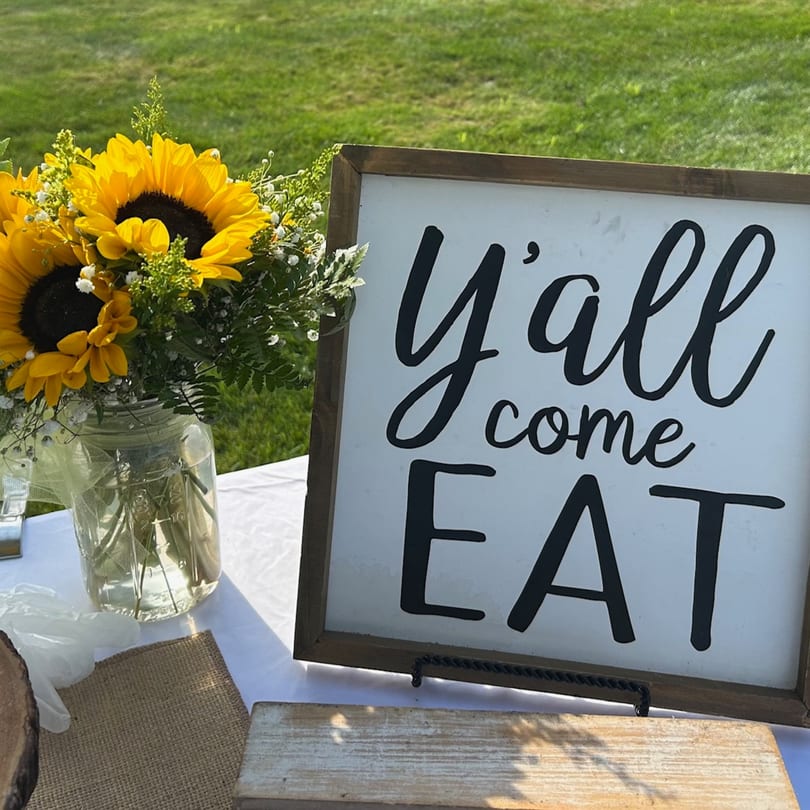 A wooden sign reading "y'all come EAT" next to a mason jar with sunflowers and greenery.