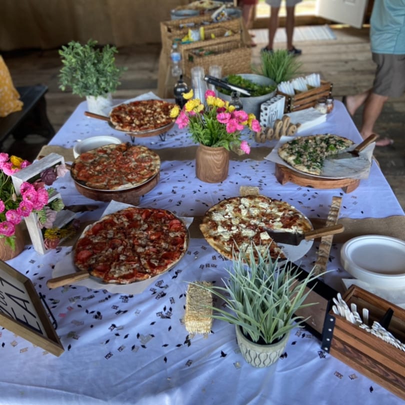A buffet table with five Marco Pizzeria pizzas, a salad bowl, and floral decorations.