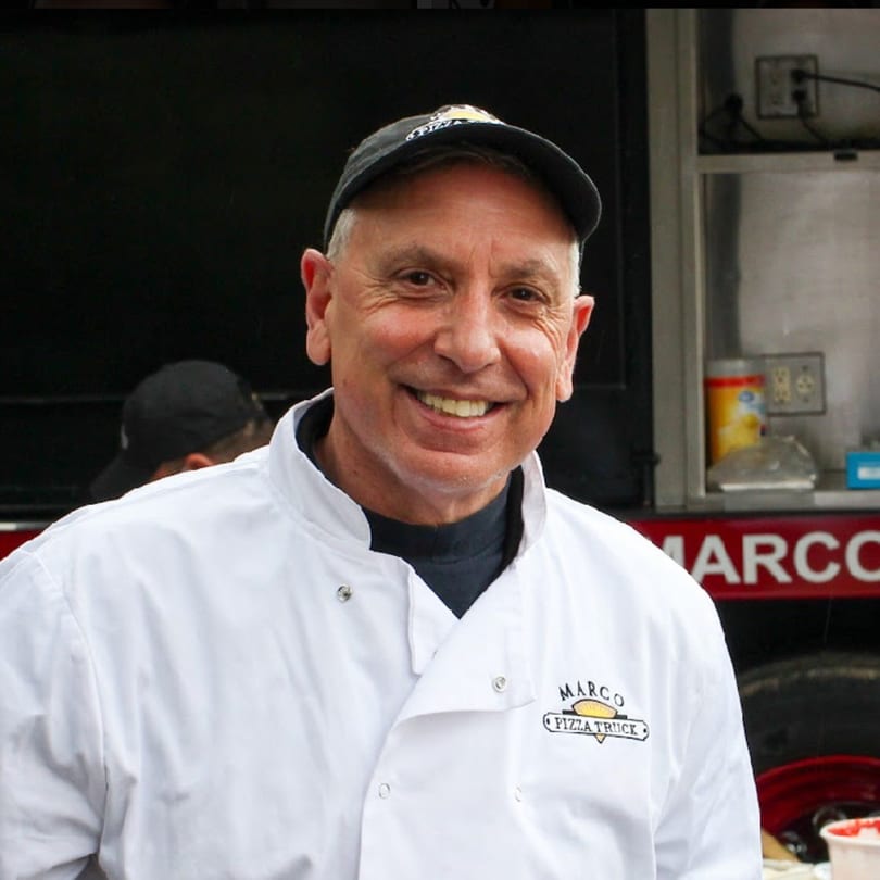 Smiling Marco Pizzeria chef in a white uniform and black cap, standing by a food truck.