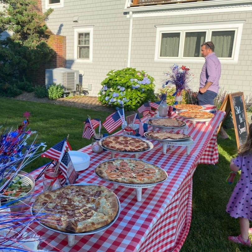 Outdoor party with pizzas on a red and white checkered table, American flags, and people.