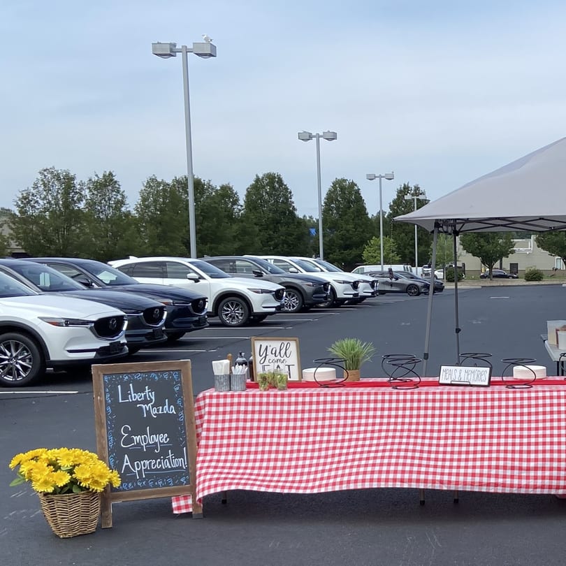 Outdoor picnic setup with red and white checkered tablecloth, signs, and cars in a parking lot.