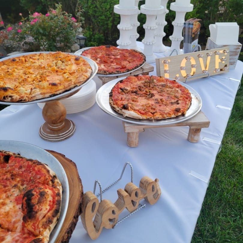 Several pizzas on stands, a "LOVE" sign, and white candlesticks on a white-covered outdoor table.